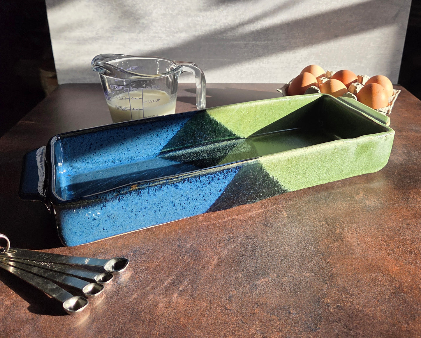 Rectangular stoneware dish in dark blue & moss glaze on a dark surface, next to a measuring cup and eggs.