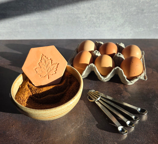 Brown sugar keeper with a maple leaf design in a bowl of brown sugar, with eggs and measuring spoons on a dark surface