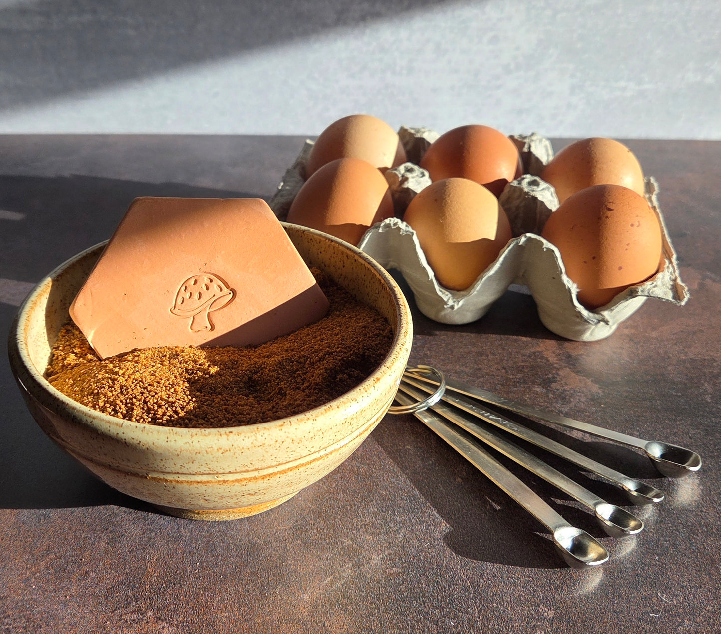Brown sugar keeper with mushroom design in a bowl of brown sugar, with measuring spoons and eggs on a brown surface