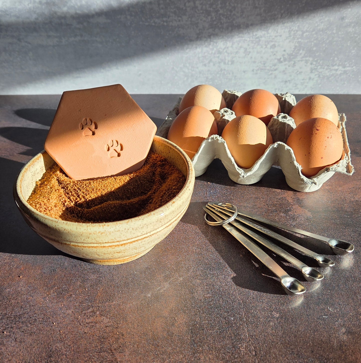 Brown sugar keeper with paw prints design in a bowl of brown sugar, carton of eggs, and metal utensils on a dark surface.