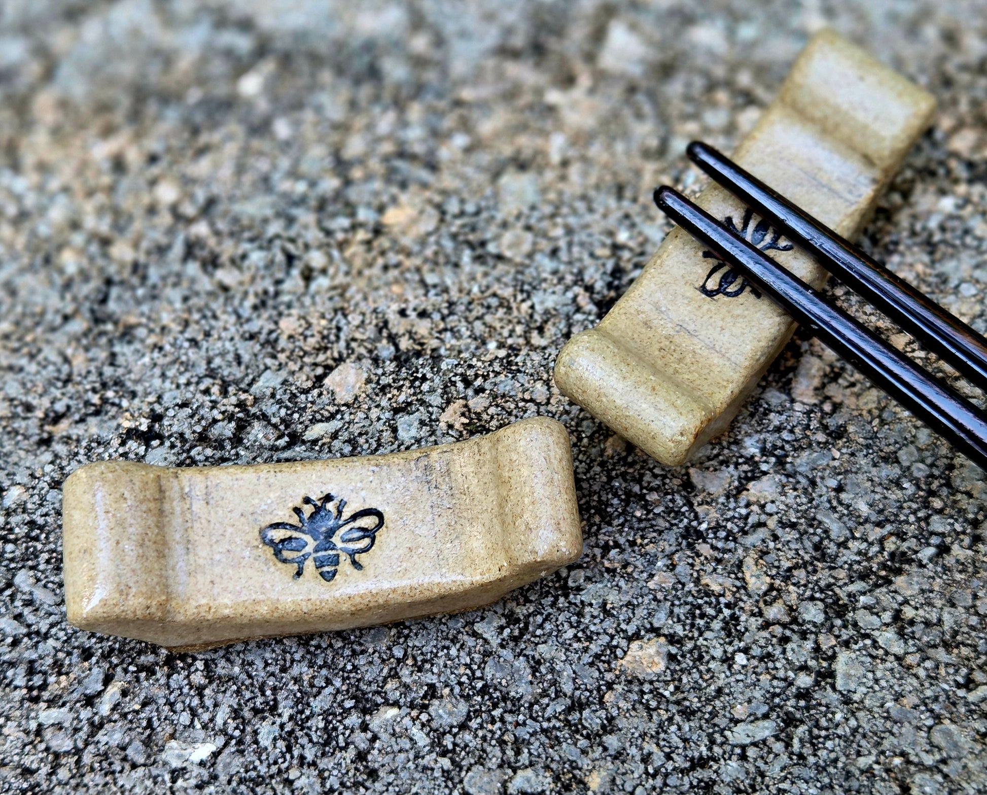 Two stoneware chopstick rests with a bee design with black chopsticks sitting on a stone surface