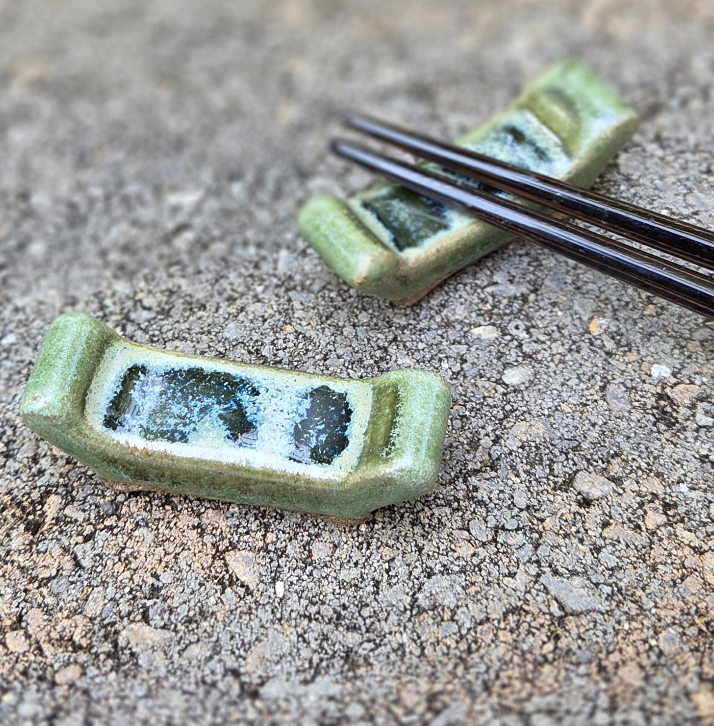Two stoneware chopstick rests with crema verde glaze with black chopsticks sitting on a stone surface
