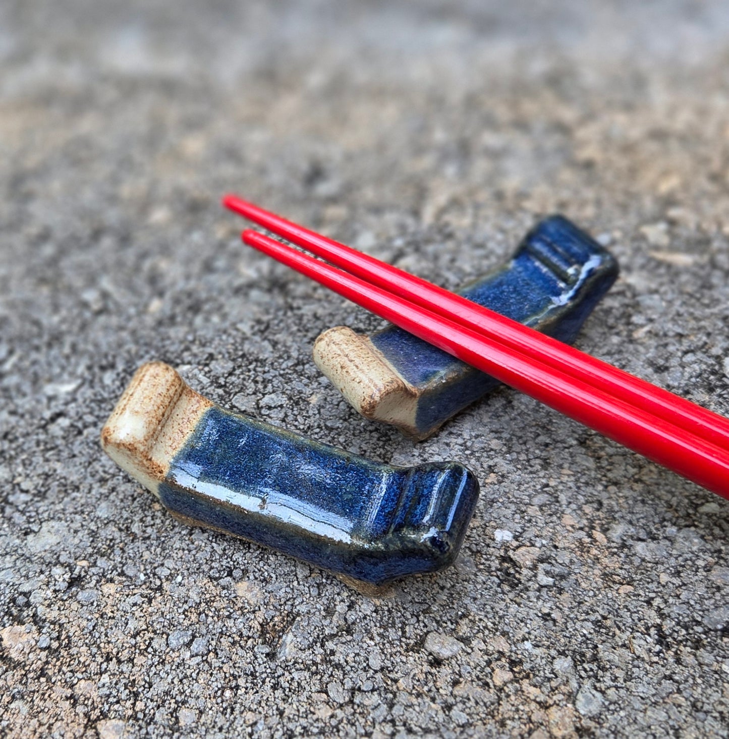Two stoneware chopstick rests with dark blue and eggshell glaze with black chopsticks sitting on a stone surface