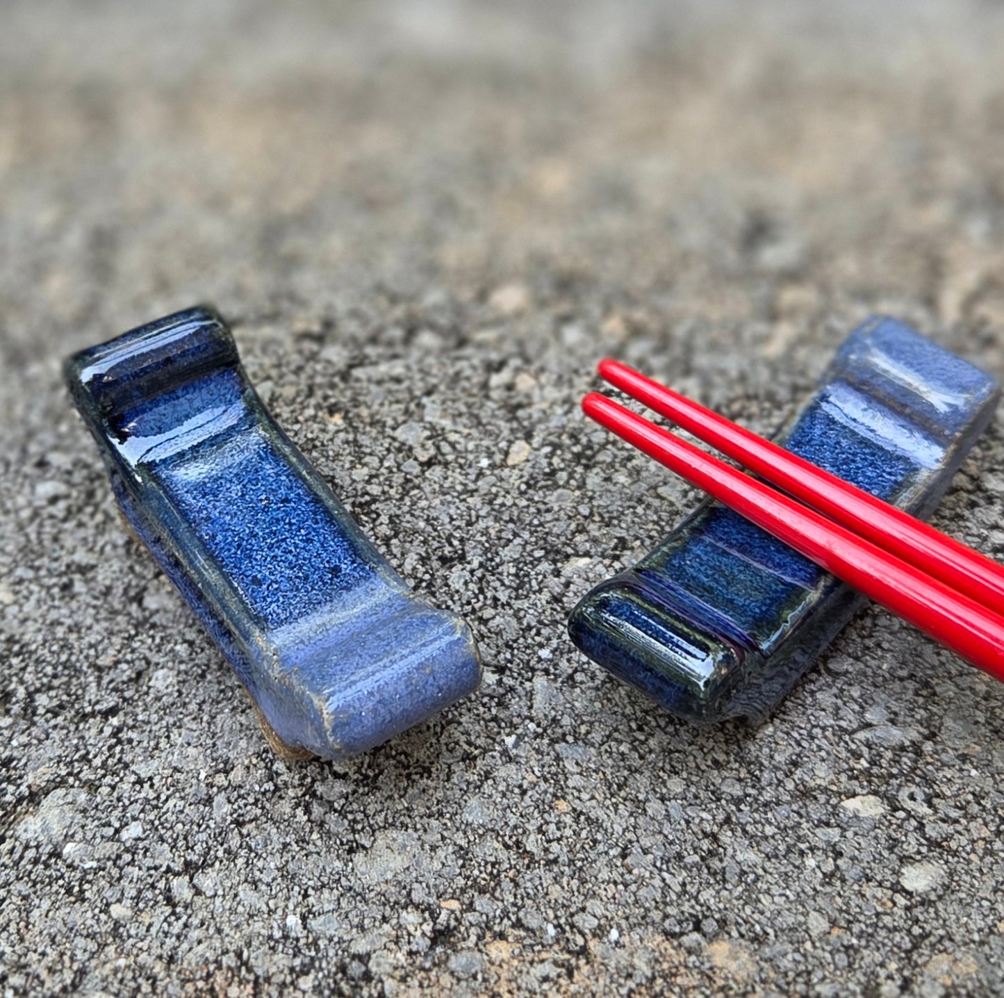 Two stoneware chopstick rests with dark blue and purple glaze with black chopsticks sitting on a stone surface