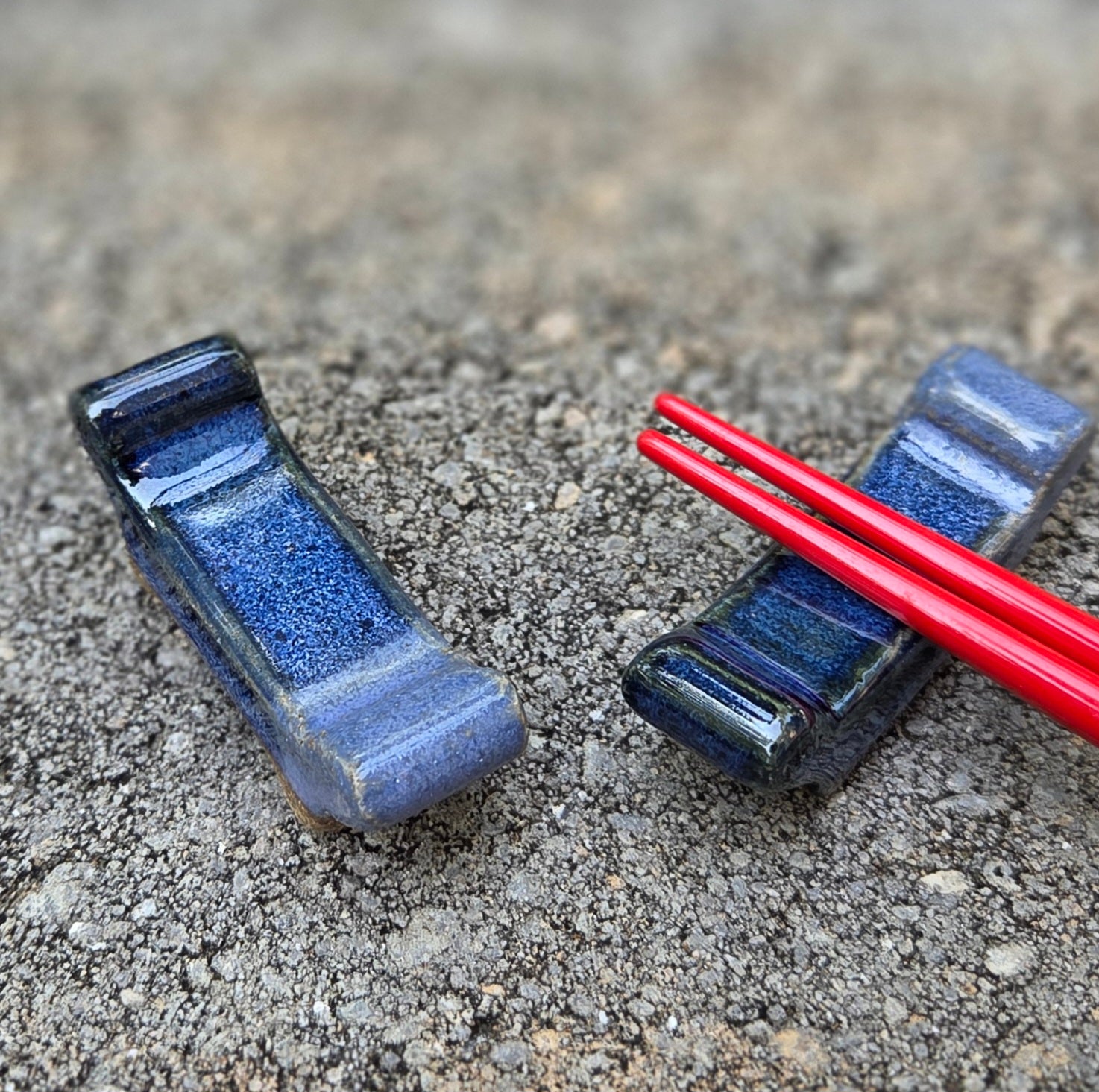 Two stoneware chopstick rests with dark blue and purple glaze with black chopsticks sitting on a stone surface