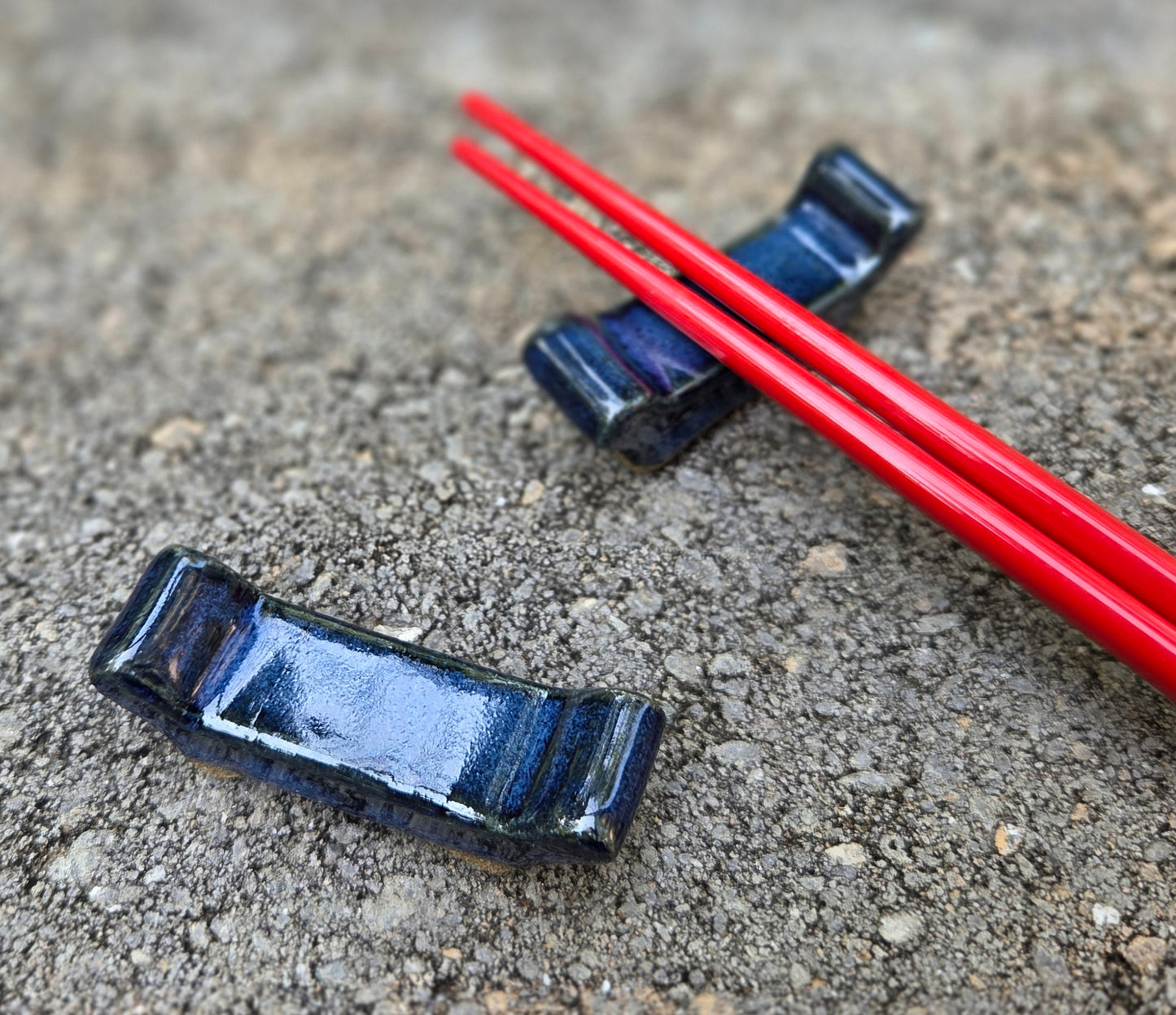 Two stoneware chopstick rests with dark blue glaze with red chopsticks sitting on a stone surface