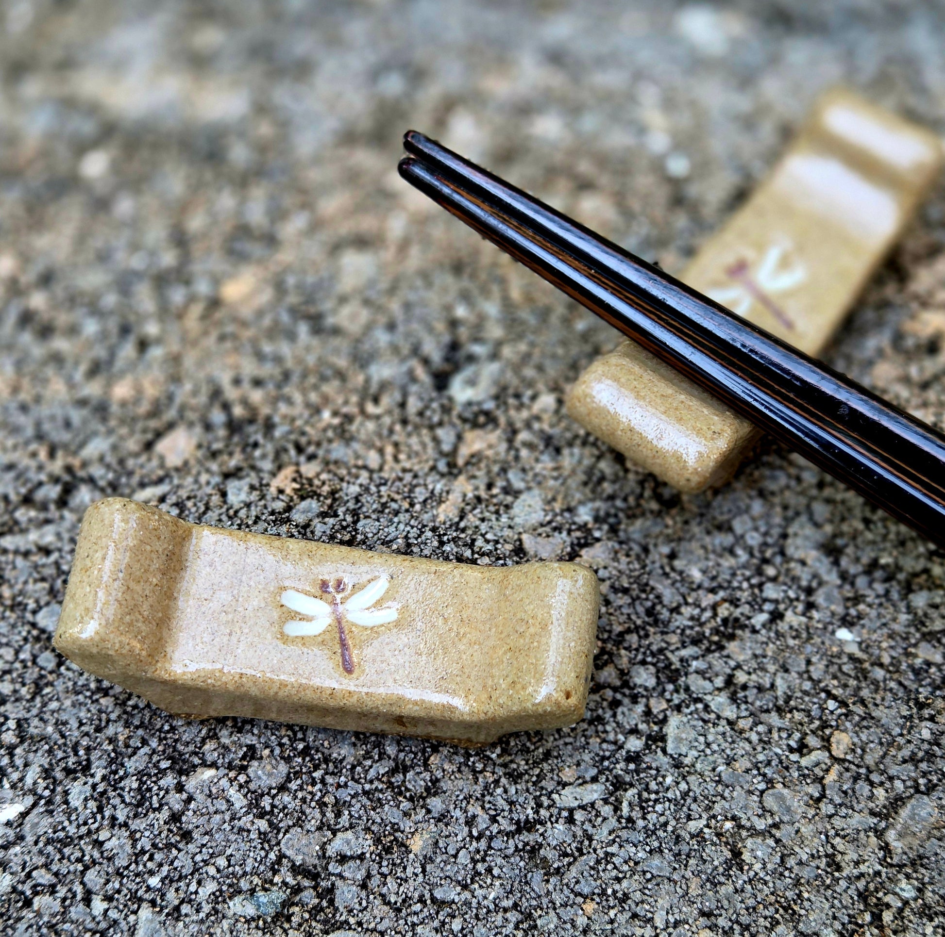 Two stoneware chopstick rests with a dragonfly design with black chopsticks sitting on a stone surface