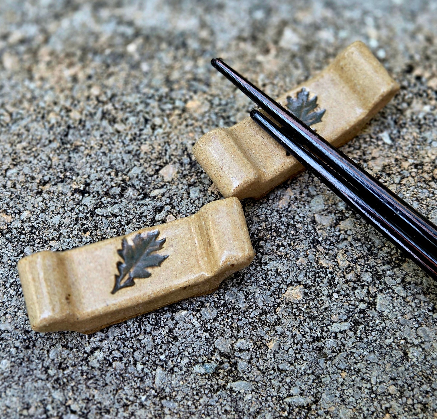 Two stoneware chopstick rests with a leaf design with black chopsticks sitting on a stone surface