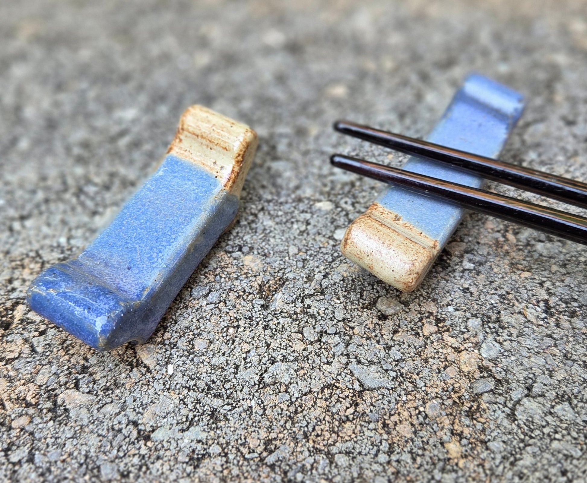 Two stoneware chopstick rests with purple and eggshell glaze with black chopsticks sitting on a stone surface