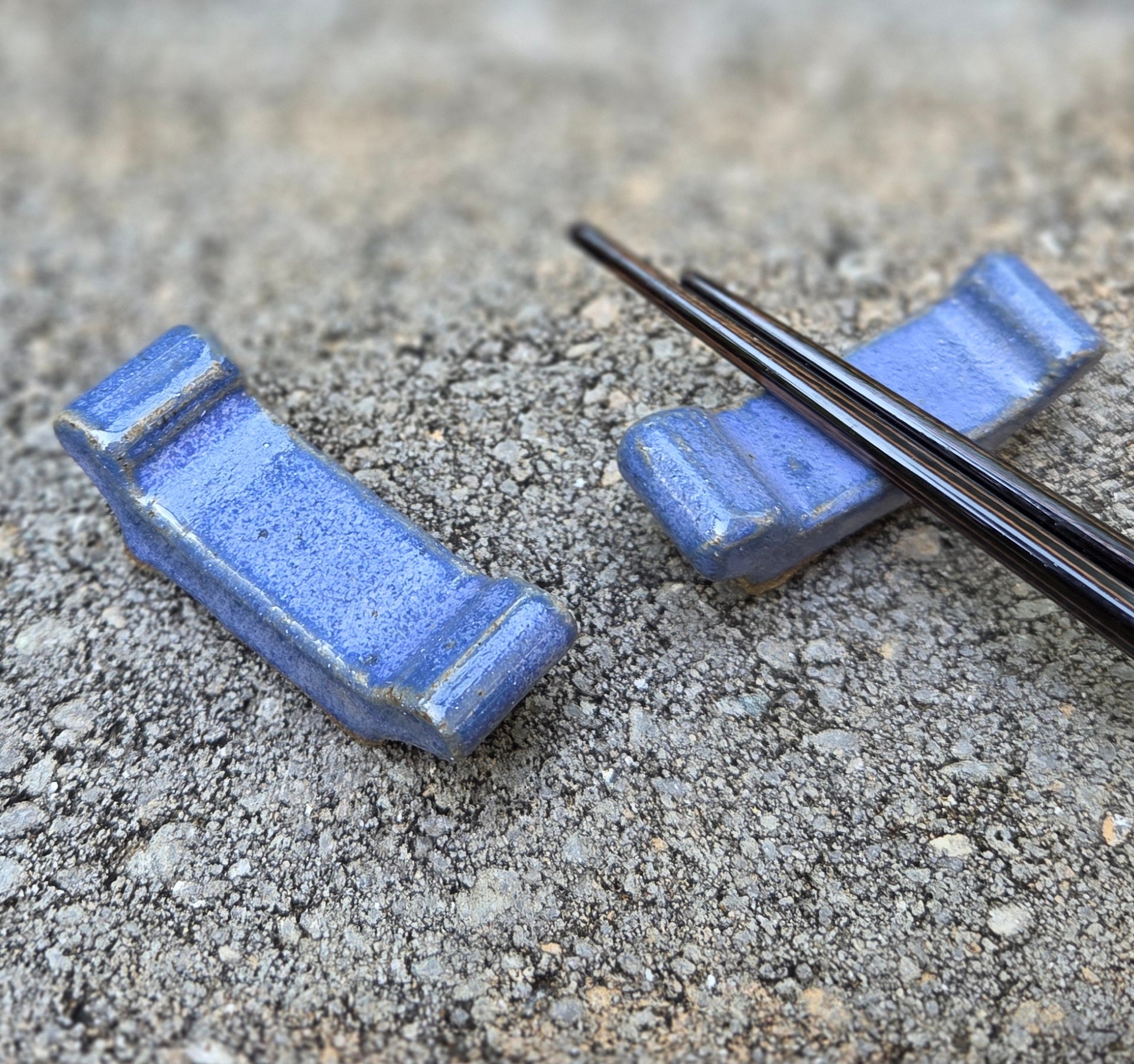 Two stoneware chopstick rests with purple glaze with black chopsticks sitting on a stone surface