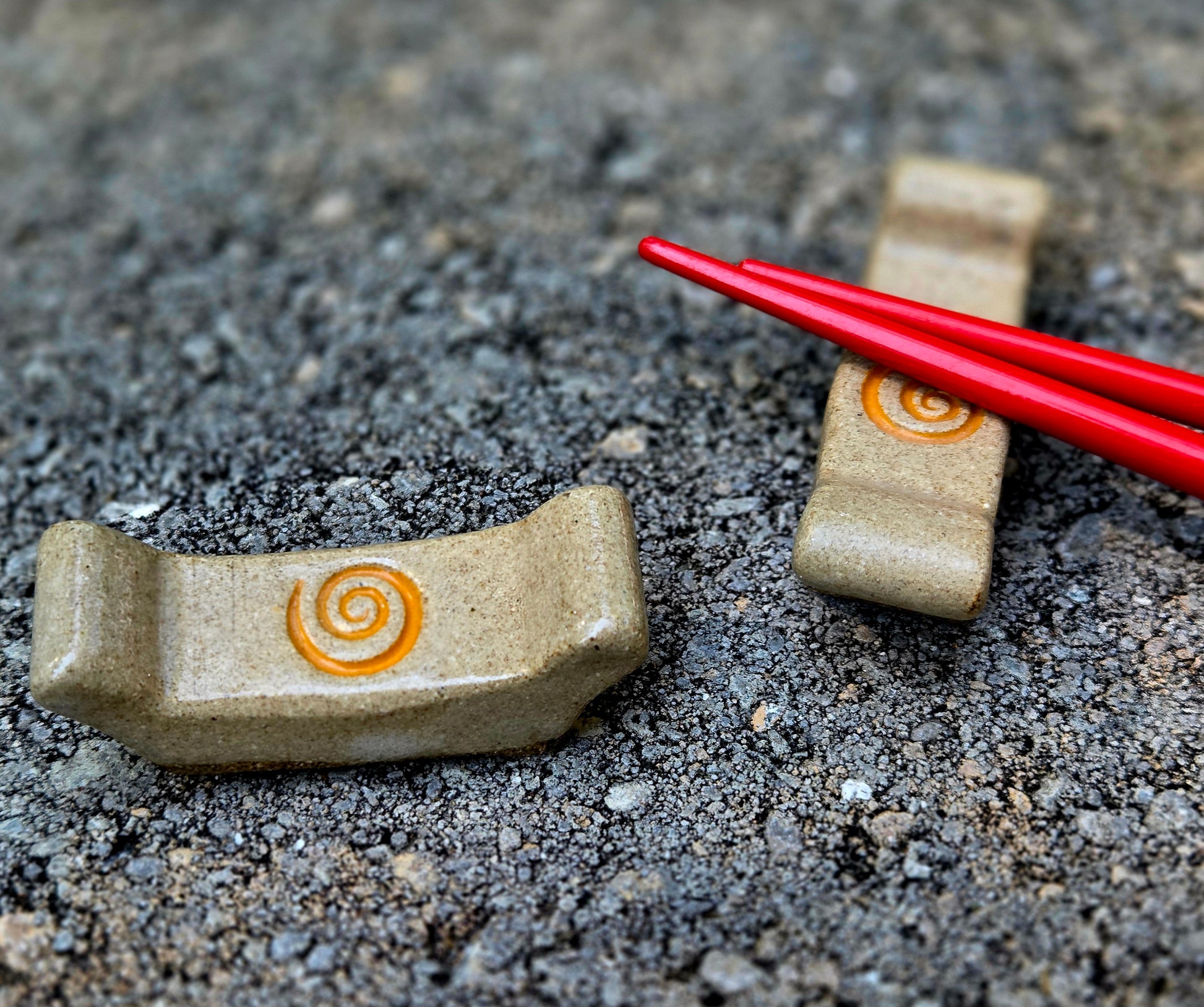 Two stoneware chopstick rests with a spiral design with red chopsticks sitting on a stone surface