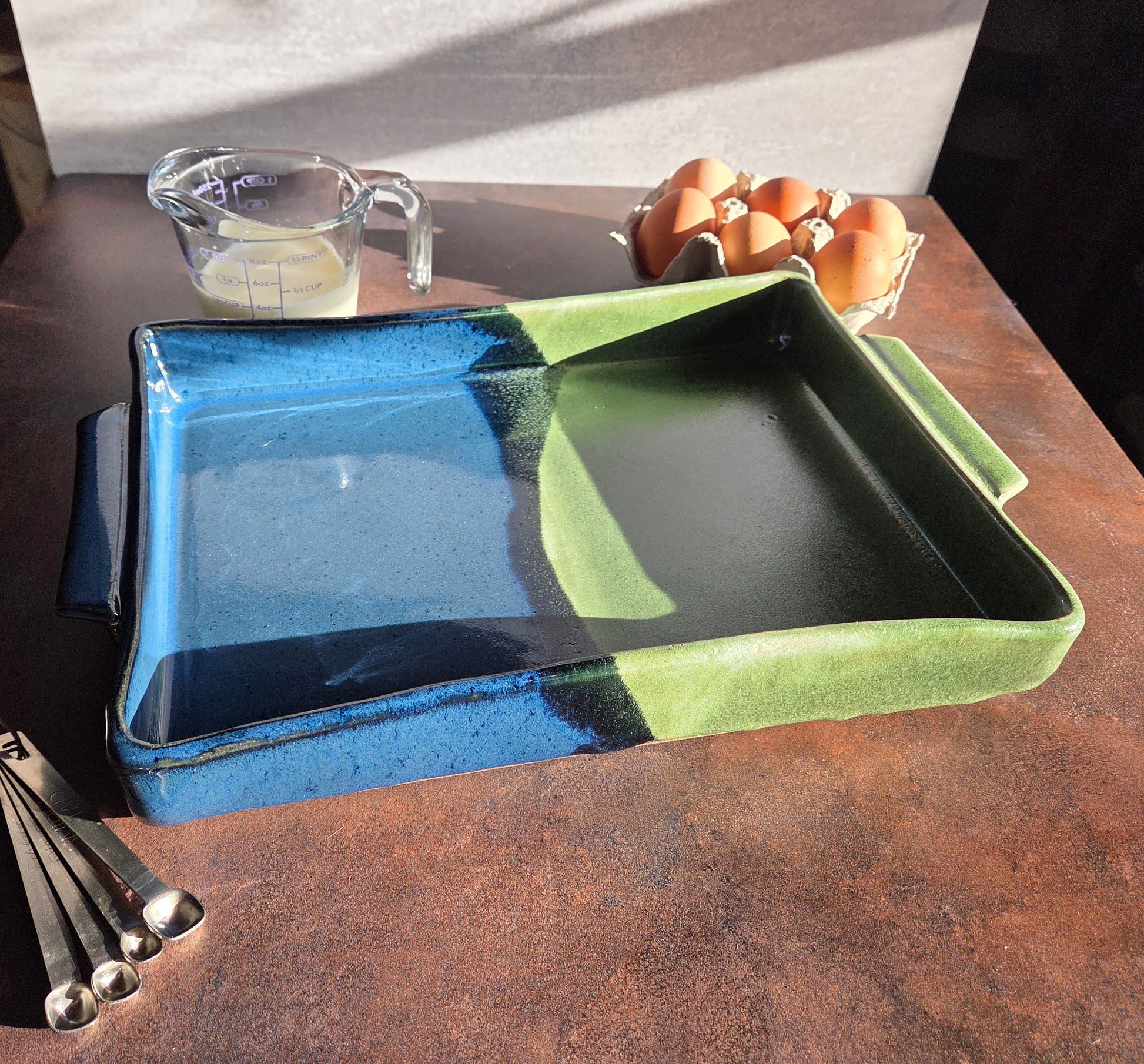 Blue and green ceramic casserole dish on a dark surface with eggs and measuring cups in the background.