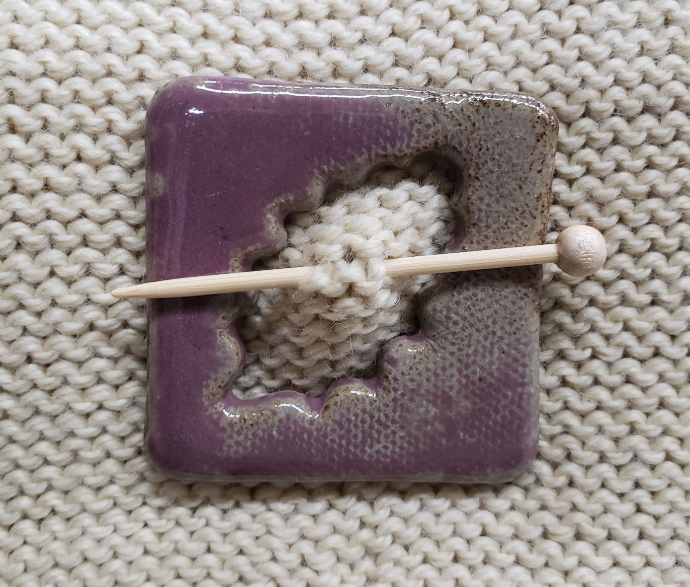 Purple ceramic shawl pin with a textured surface attached to beige knit fabric